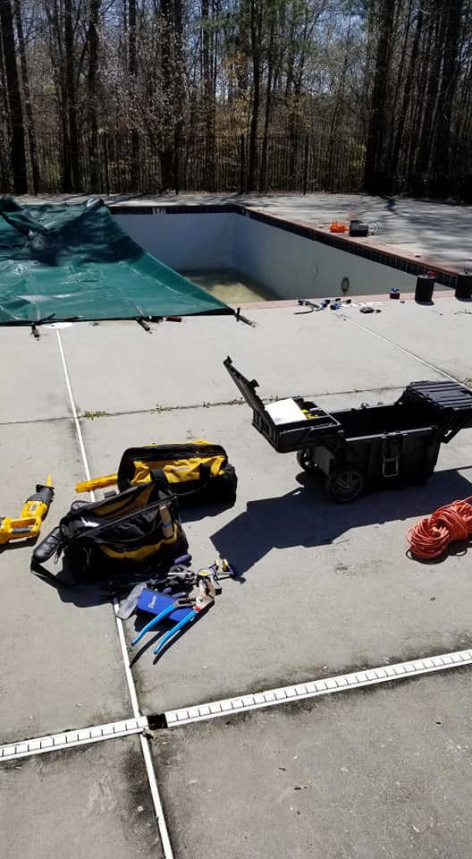 Pool renovation tools and equipment on concrete deck next to a covered, empty swimming pool, surrounded by trees, illustrating maintenance and upgrade services for aquatic facilities.