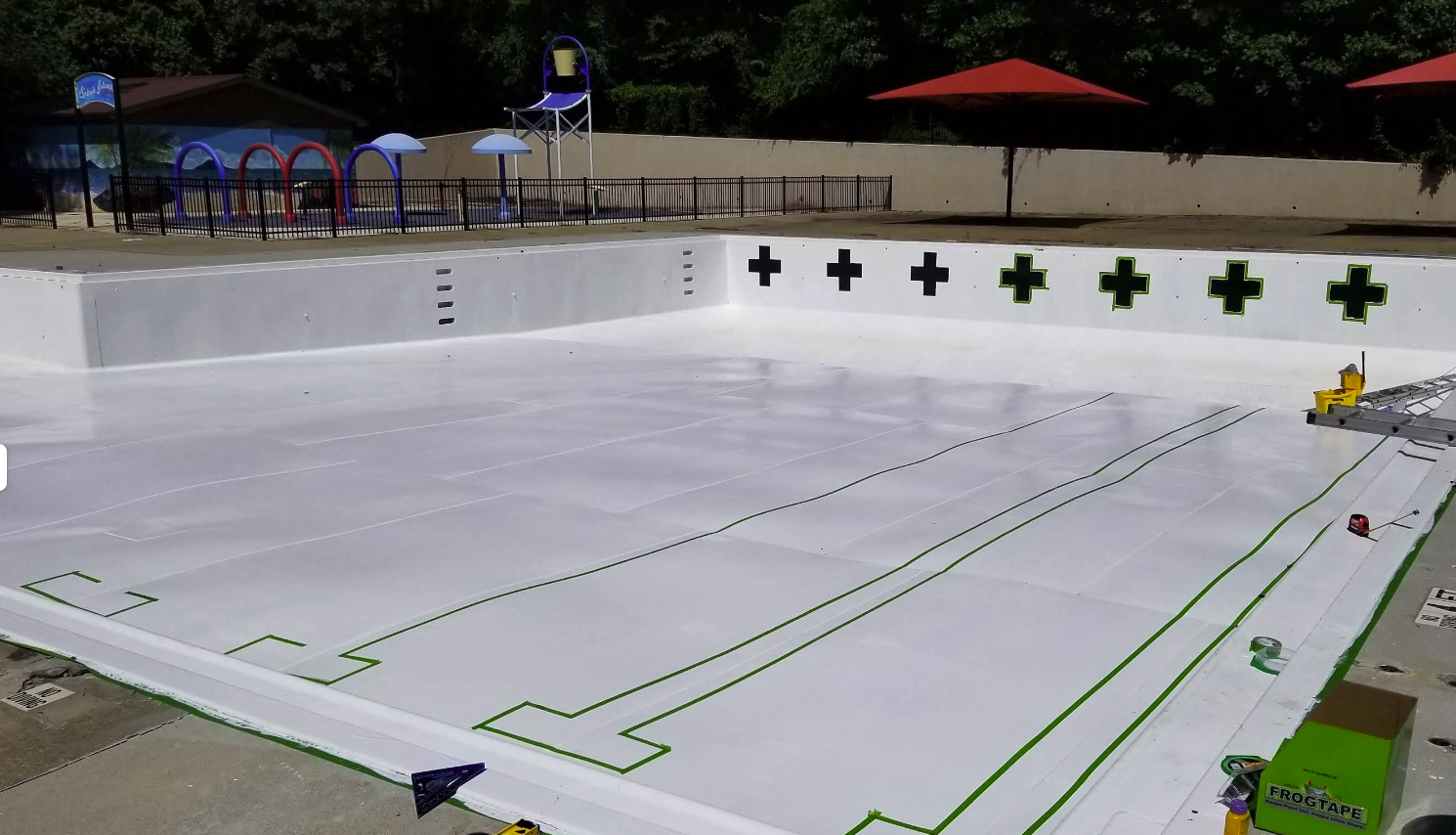 Pool renovation in progress, showcasing a partially filled swimming pool with white surface and green tape outlines, surrounded by umbrellas and a water slide in a commercial facility.