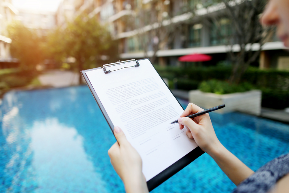 Person reviewing a pool management contract by a swimming pool, emphasizing pool auditing and compliance services.