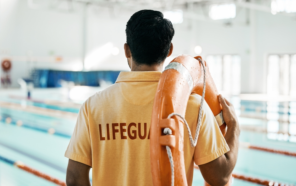 Lifeguard in yellow shirt with "LIFEGUARD" text, holding an orange life preserver, overseeing an indoor swimming pool environment, emphasizing safety and compliance in aquatic facilities.