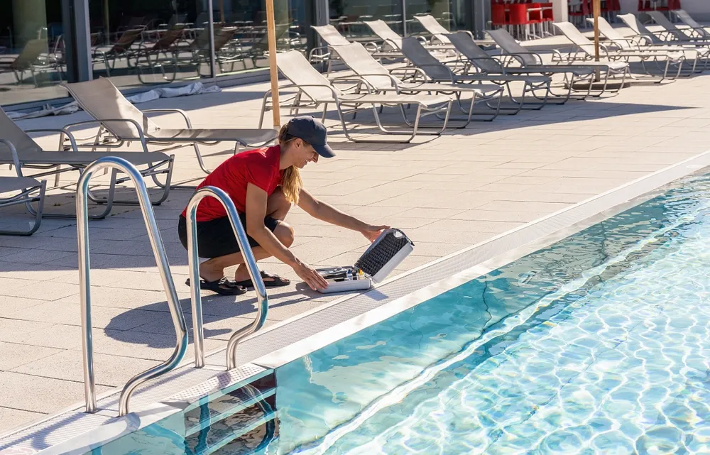 Pool maintenance professional inspecting water quality with testing kit by the poolside, surrounded by lounge chairs, emphasizing the importance of routine maintenance for aquatic facility safety.