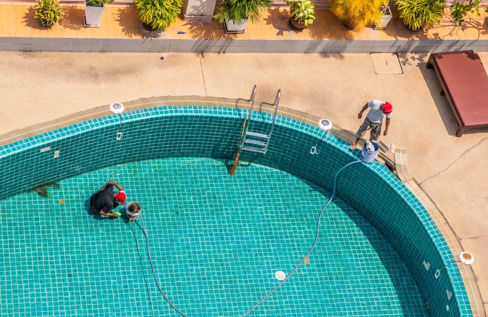 Pool maintenance workers cleaning a swimming pool with a vacuum and tools, surrounded by potted plants, emphasizing the importance of water quality management and safety in aquatic facilities.