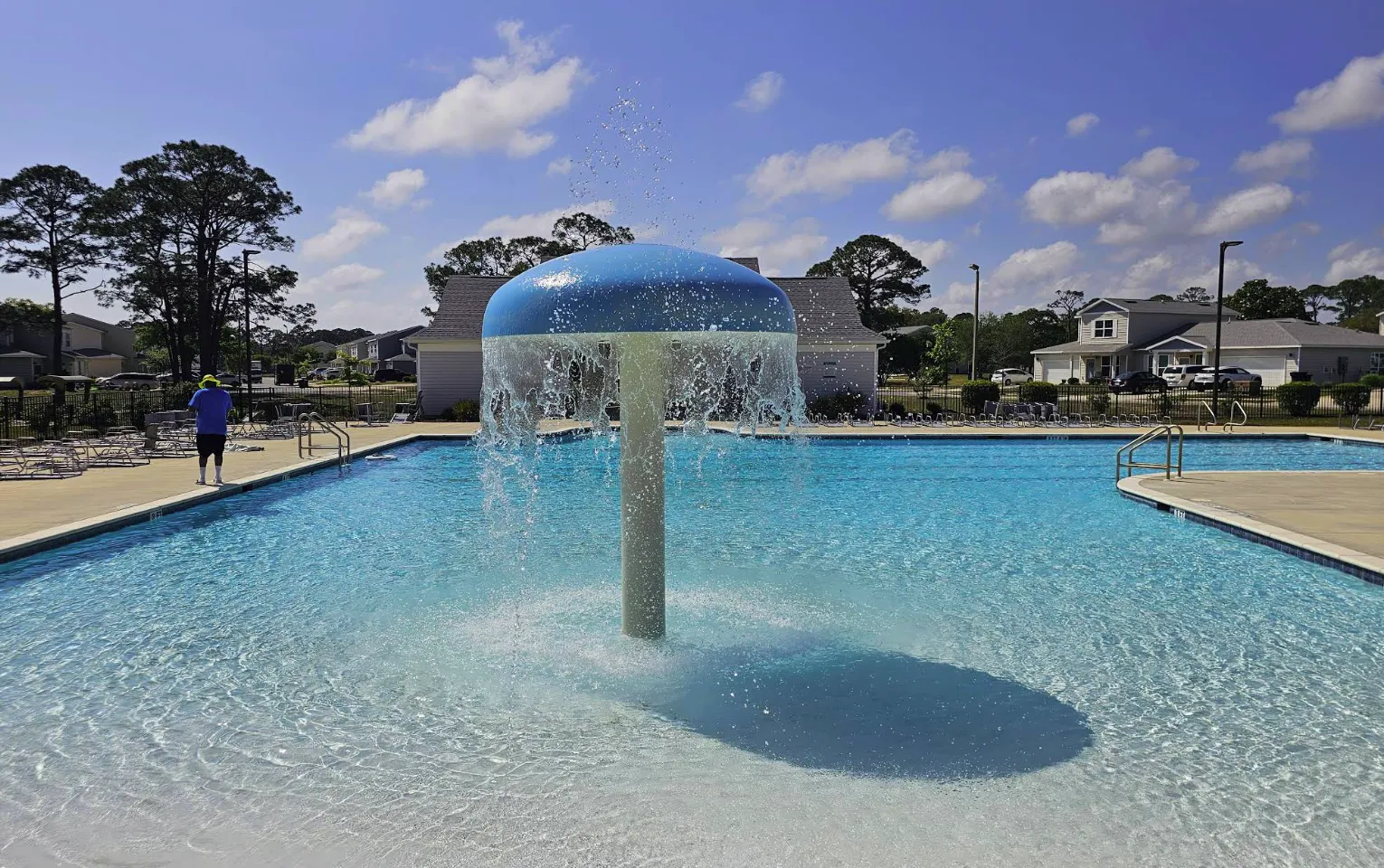 Pool with a blue mushroom fountain spraying water, surrounded by clear water and lounge chairs, with a person in a blue shirt walking nearby, illustrating a recreational setting for spring pool maintenance and enjoyment.